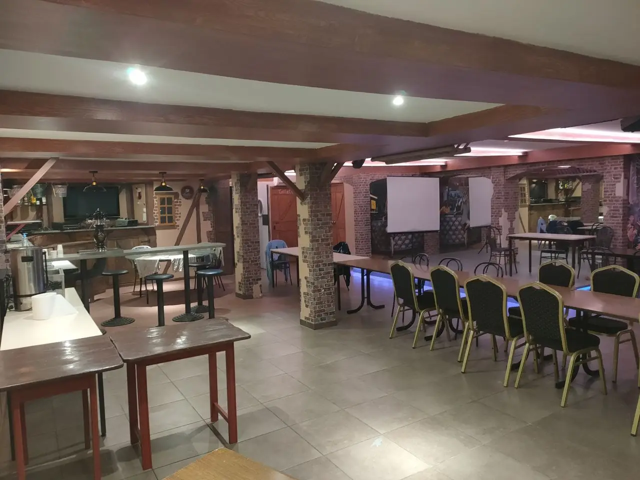 Salle de réception de Mont Relais Gaillard, avec tables, poutres en bois, grand écran et espace bar.